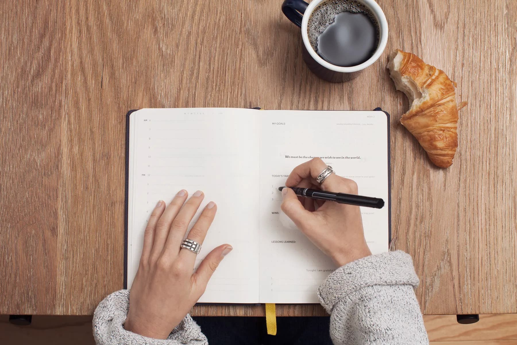 Hands writing in a planner next to a cup of coffee on a wooden table